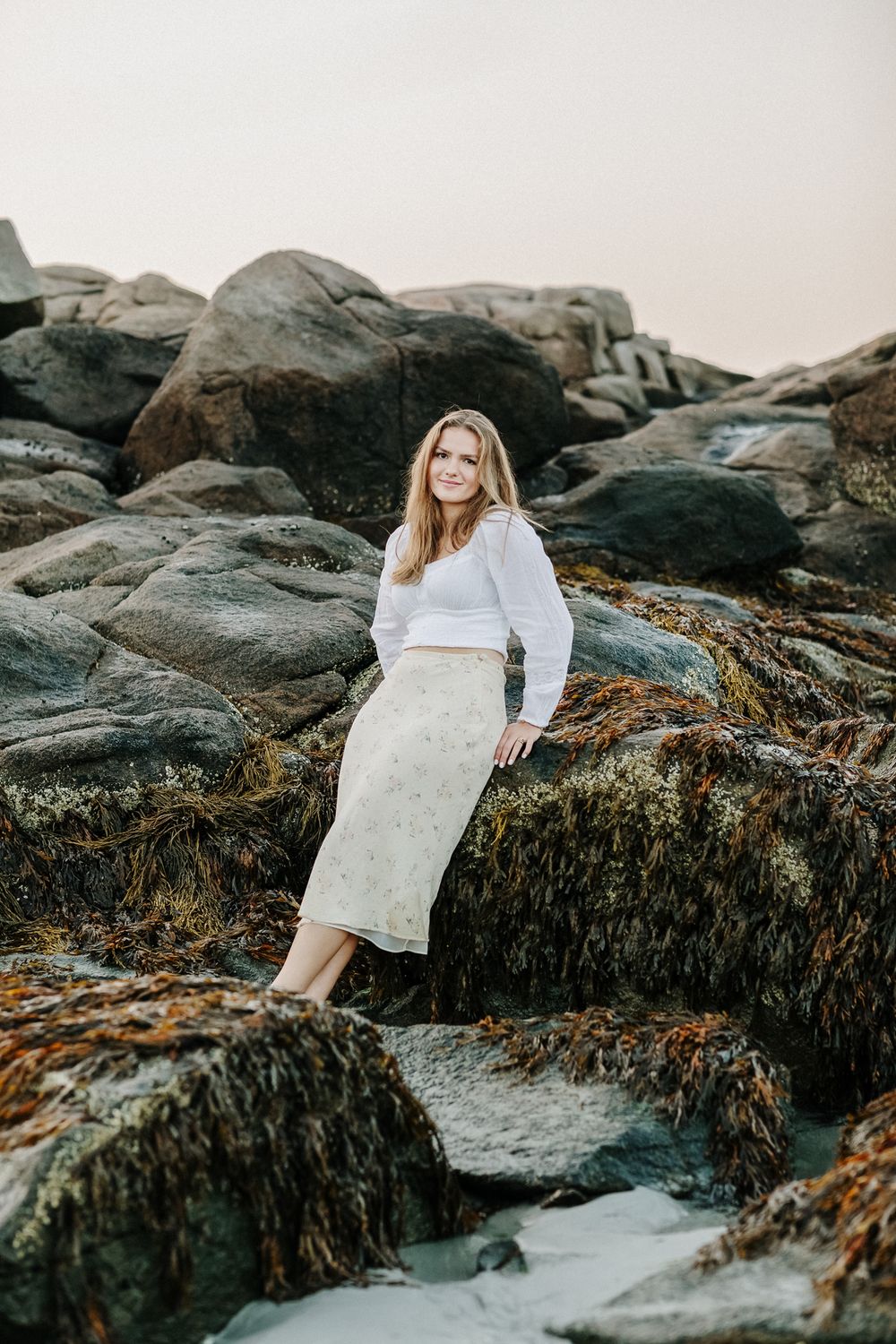 Blond senior girl wearing a pale yellow floral skirt leans back on dark coastal rocks at Wingaersheek Beach in Gloucester for senior photos.