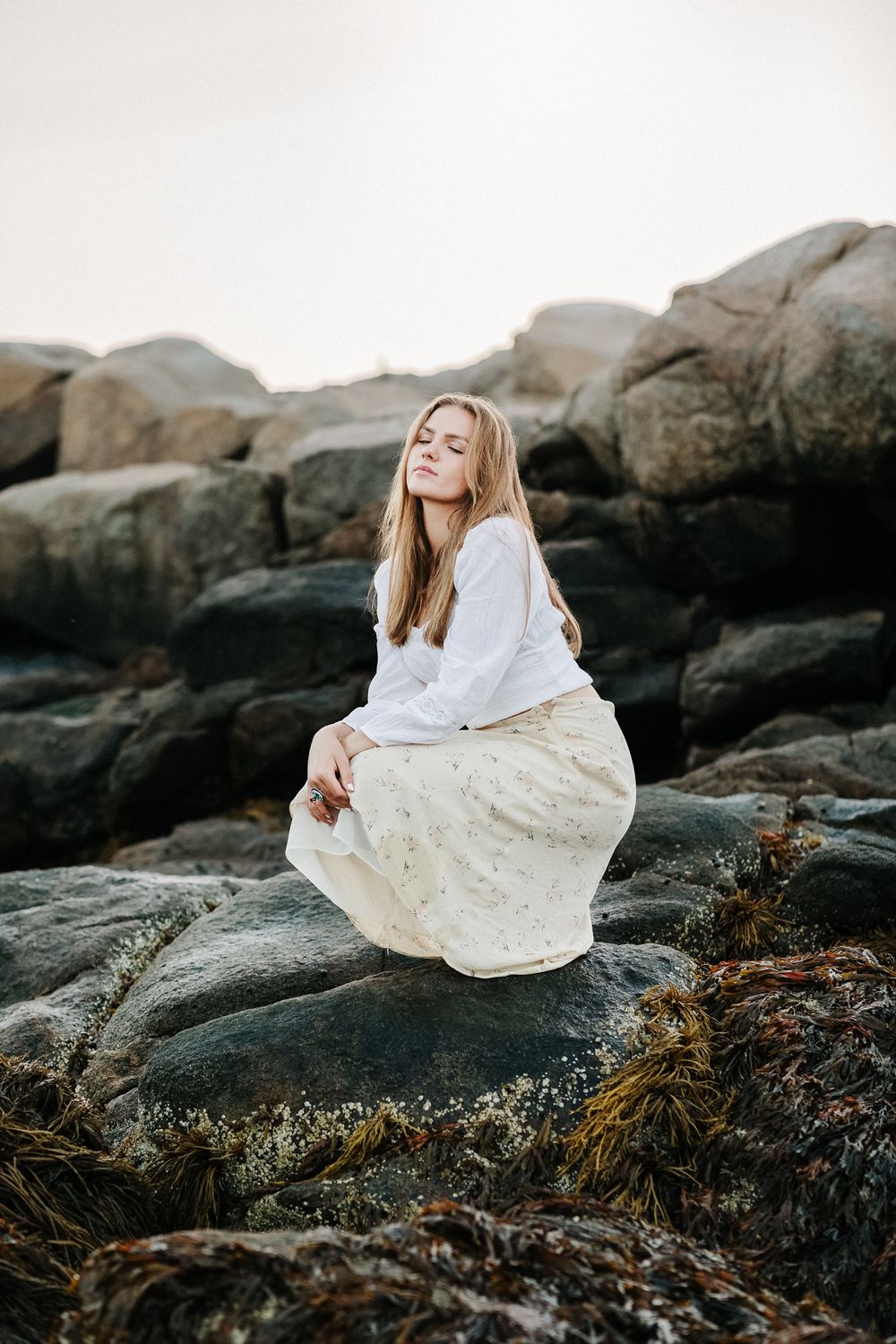 Blond senior girl poses serenely with eyes closed on dark coastal rocks at Wingaersheek Beach in the North Shore Gloucester, MA.