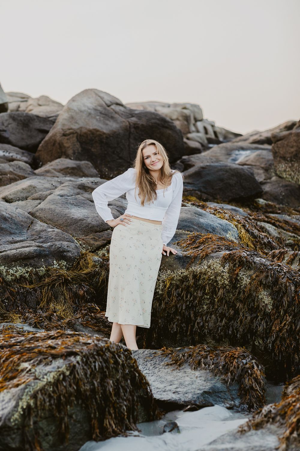 Blond senior girl poses with hand on hip leaning on dark coastal rocks for senior photos at Wingaersheek Beach Gloucester, MA, North Shore.