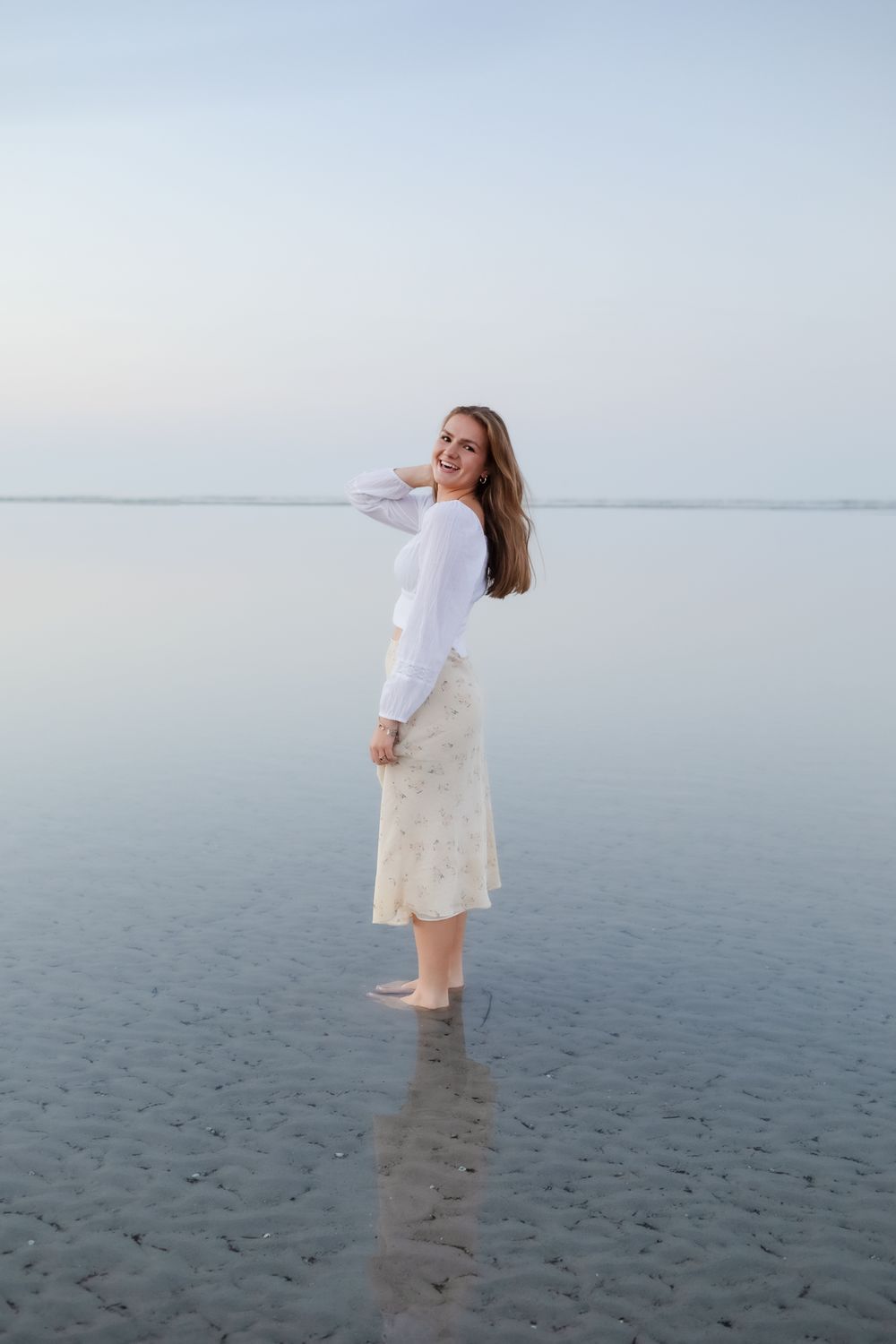 Blond girl wears a white blouse and pale yellow skirt posing in shallow blue ocean water for senior photos at Wingaersheek Beach.