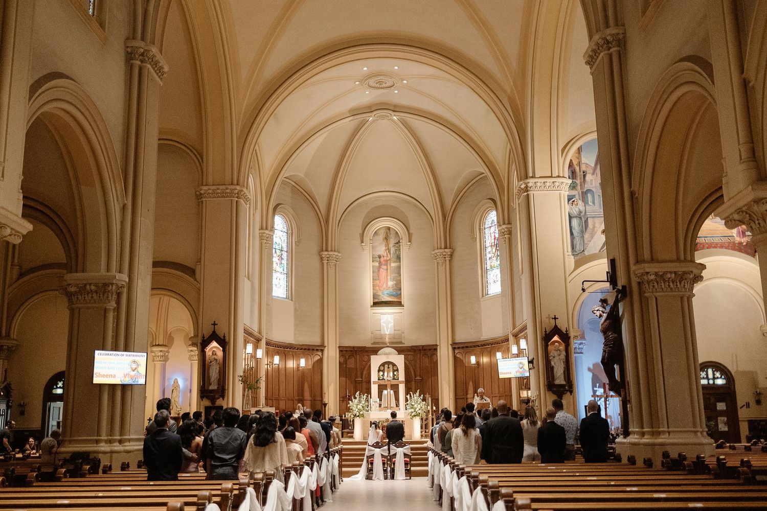 Elegant Catholic church wedding ceremony with guests seated in wooden pews beneath arched ceilings and stained glass windows.