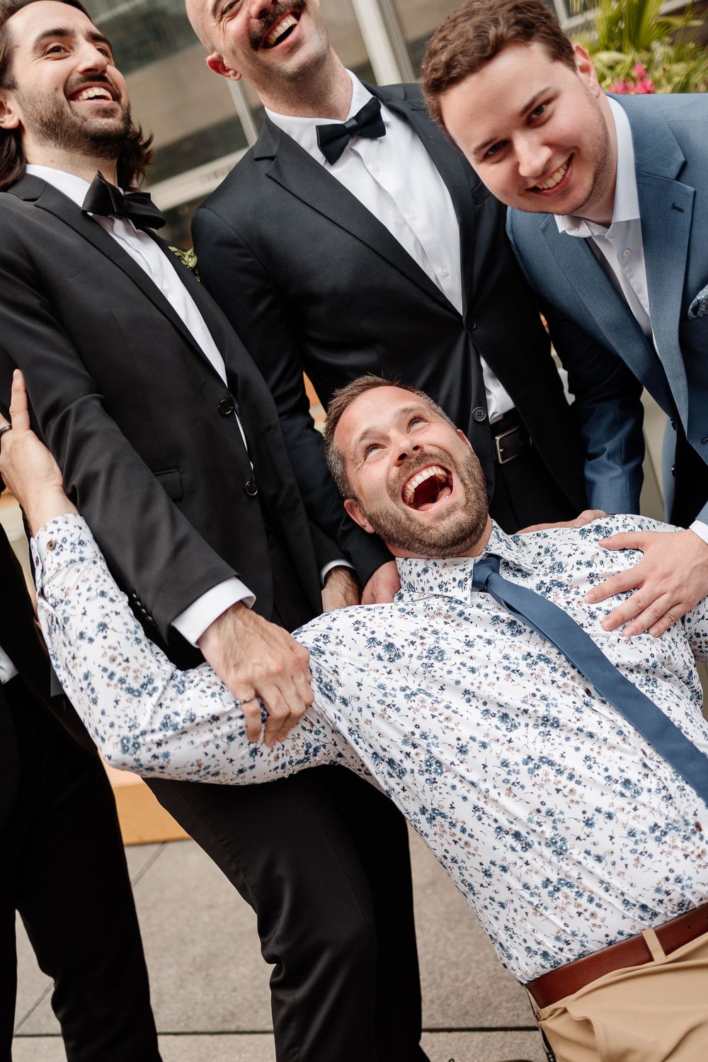 Groomsmen in black tuxedos laughing and playfully lifting a man in a floral shirt at a wedding.