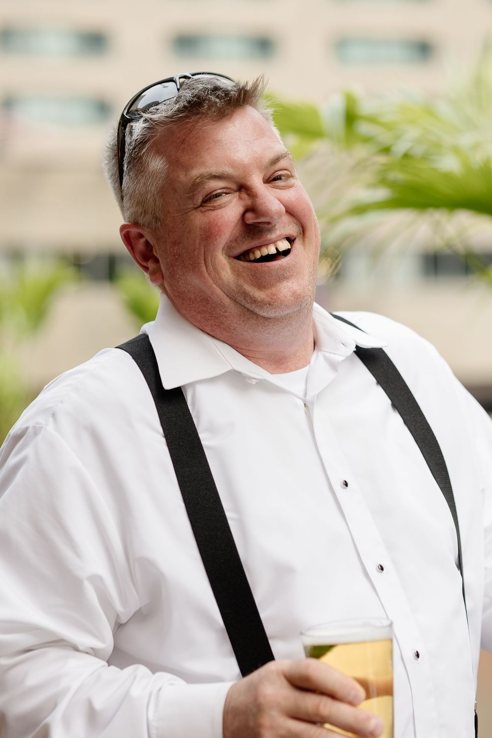Cheerful middle-aged man in white shirt with suspenders laughing outdoors while holding a glass of beer. Candid wedding photography Montreal