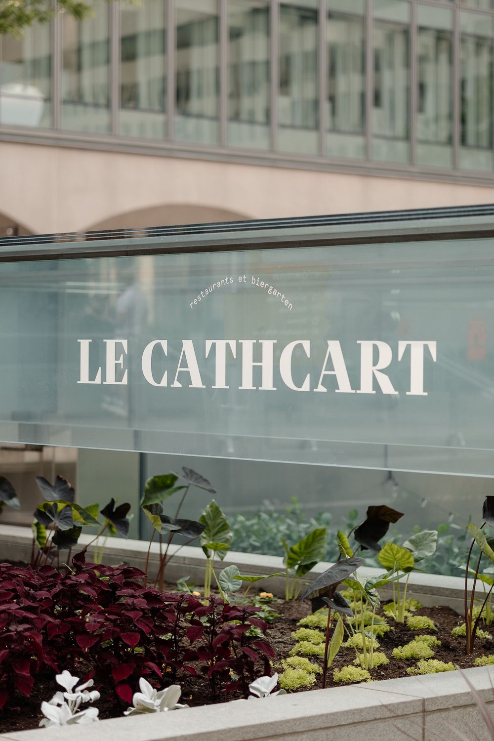 Le Cathcart wedding venue sign on glass facade surrounded by lush green and burgundy plants in in downtown Montreal.