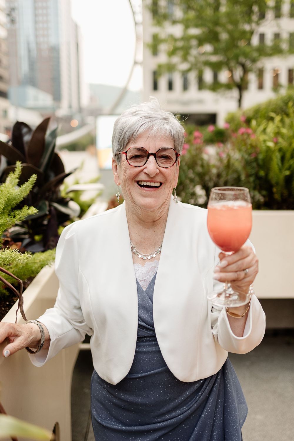 Candid photo of a grandma with short gray hair and glasses raising a pink cocktail glass outdoors at Le Cathcart Wedding Venue.