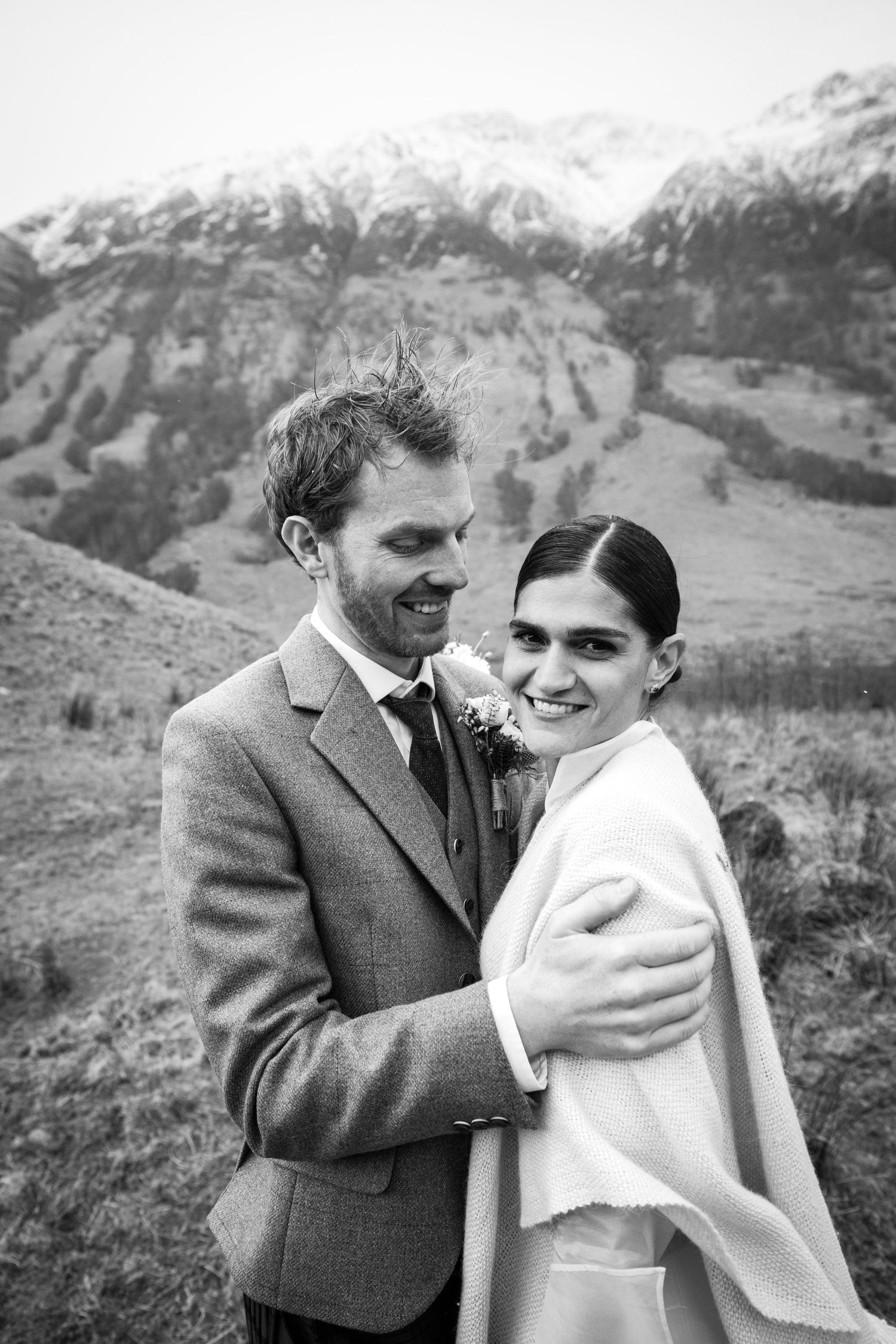 Black and white photo of a smiling bride and groom embracing with snow-capped mountains in the background.