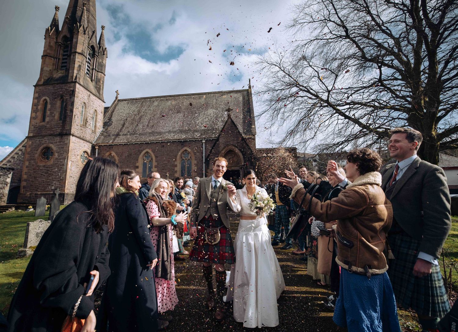 Bride and groom exit a historic stone church surrounded by cheering guests throwing confetti on a bright winter wedding day.