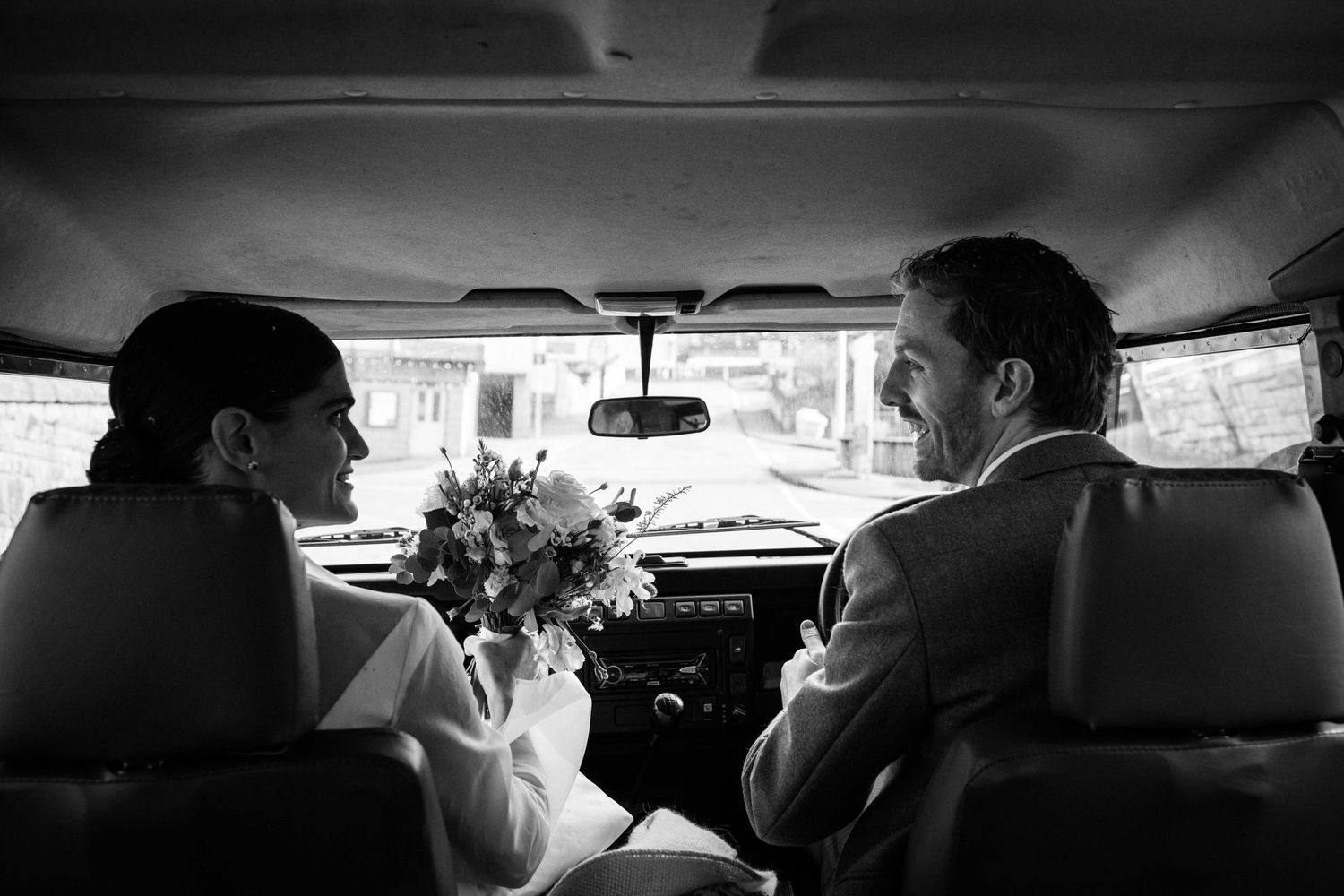 Black and white photo of a bride and groom sitting in a vintage car, bride holding a bouquet, sharing an intimate moment.