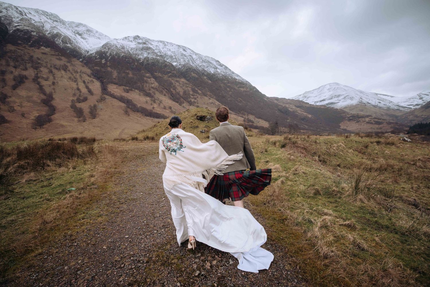 Couple in wedding attire walking hand-in-hand on a gravel path toward snow-capped Scottish Highland mountains.