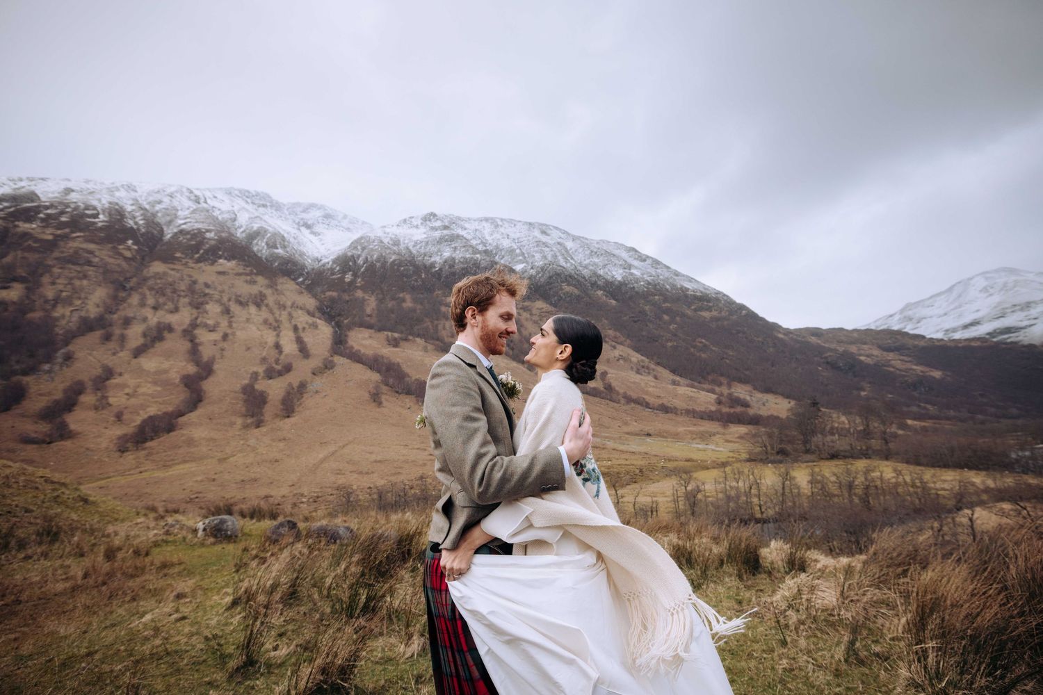 Couple embracing in Scottish Highlands with snow-capped mountains, groom in kilt, bride in white dress.