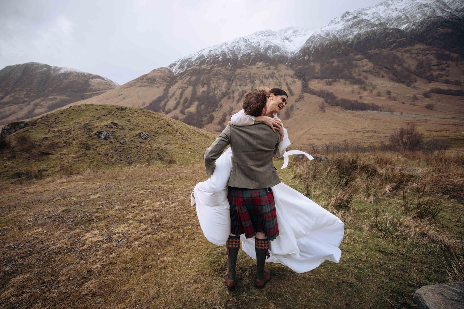 Groom in tartan kilt lifts bride in white dress outdoors with misty Scottish Highland mountains in background.