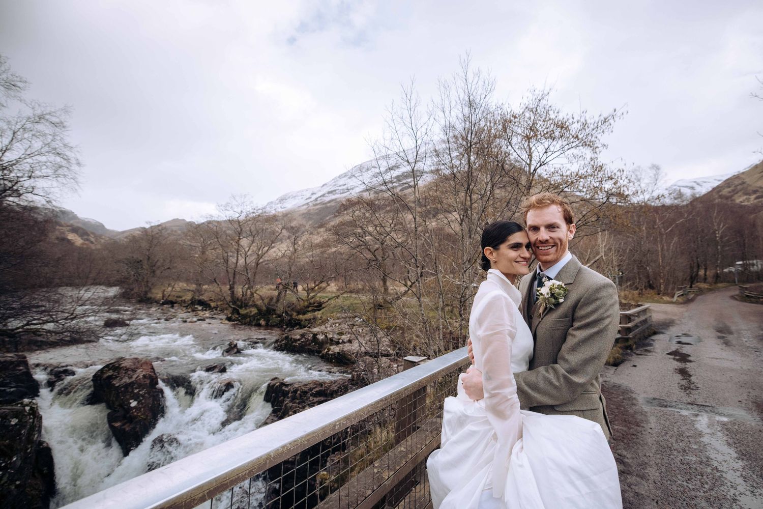 Bride and groom smiling on a bridge with a rushing river and misty mountain backdrop on their wedding day.