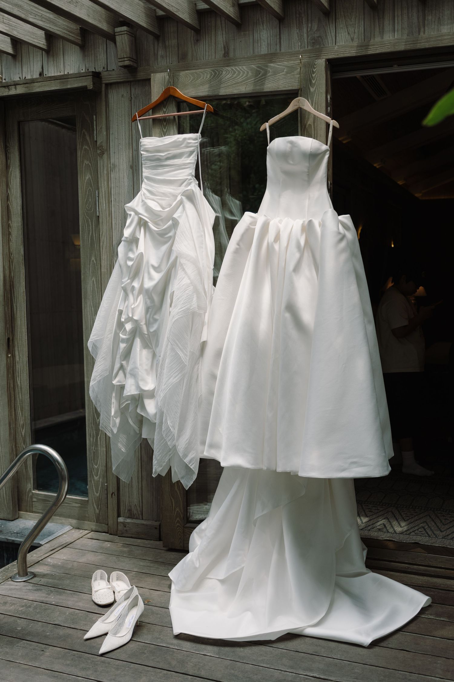 Two white wedding gowns hanging on wooden posts outdoors with white bridal heels below.
