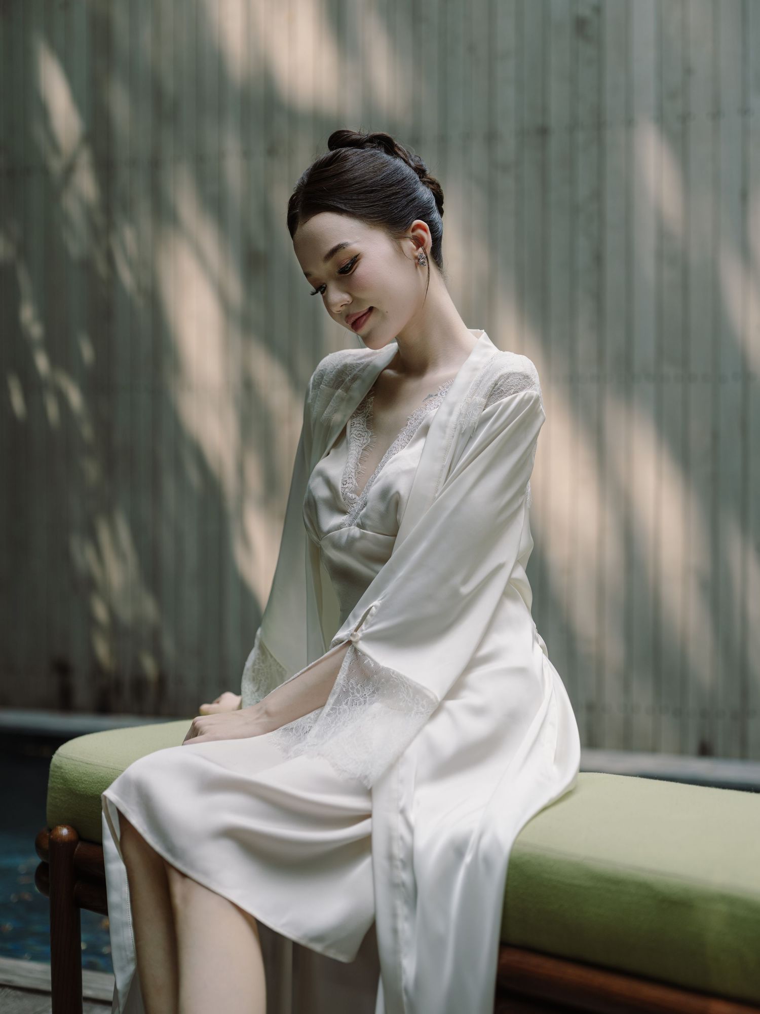 Elegant Asian woman in white silk robe and lace slip dress seated gracefully in a serene outdoor setting.