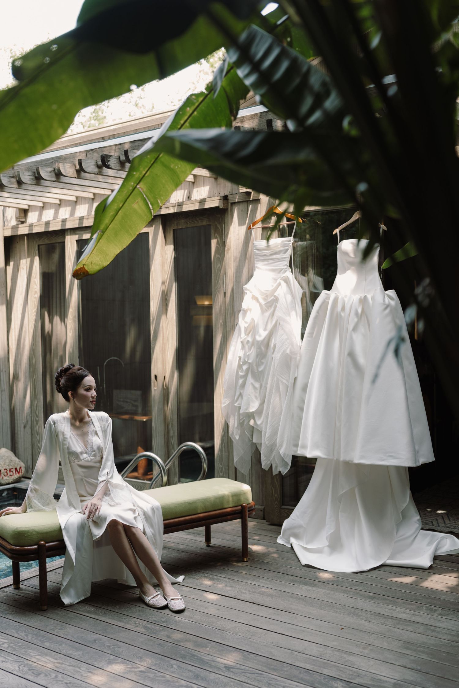 Bride in white robe sitting on chaise lounge on wooden deck beside hanging wedding gowns surrounded by tropical plants.