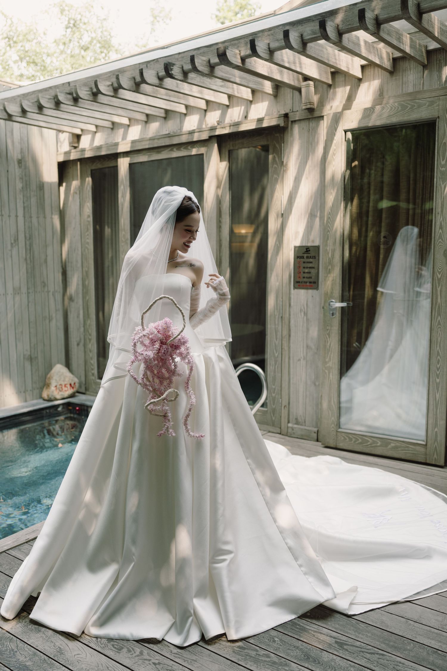 Bride in strapless ball gown with cathedral veil holds pink bouquet on wooden deck beside pool.