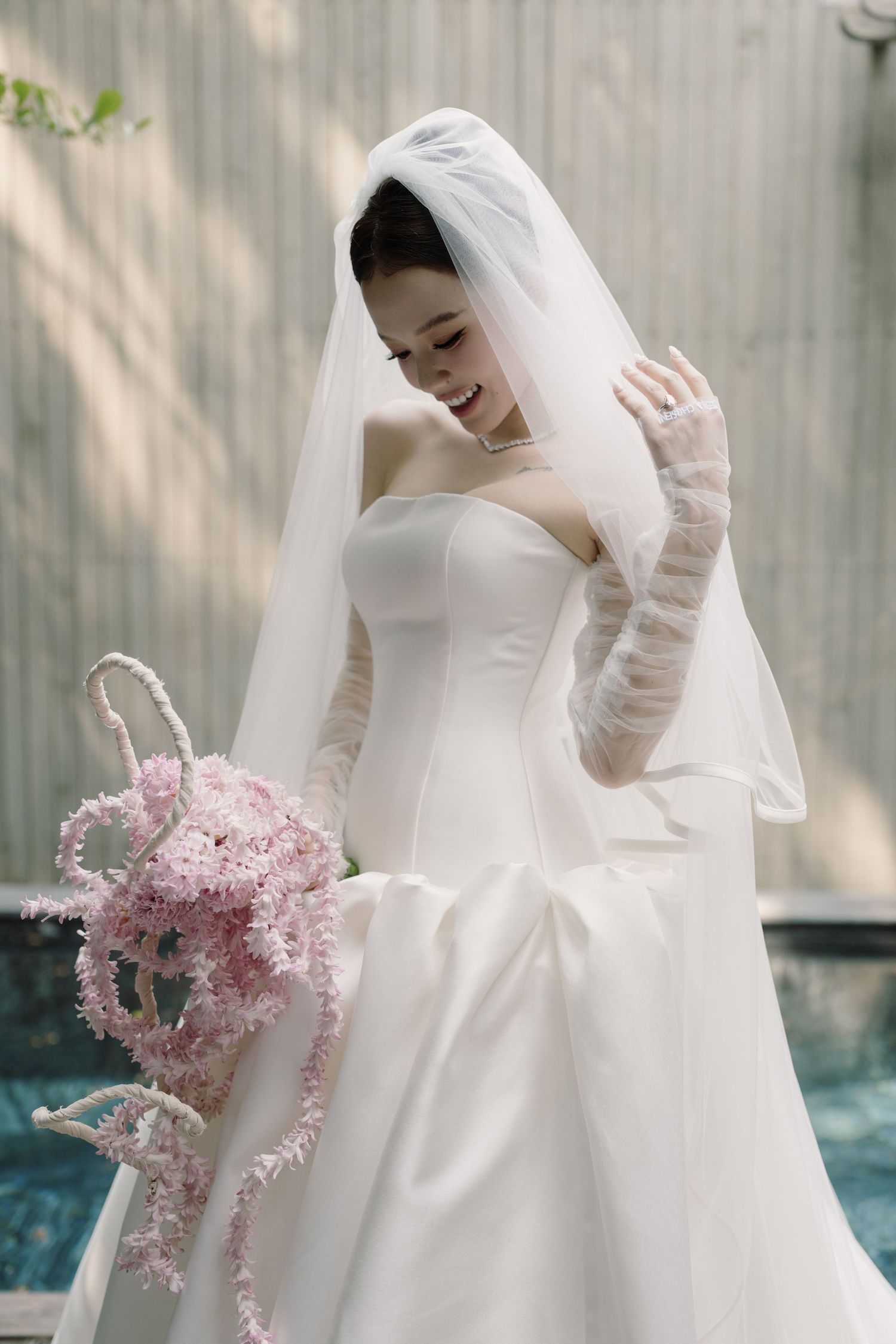 Smiling bride in white strapless gown holding pink floral bouquet.