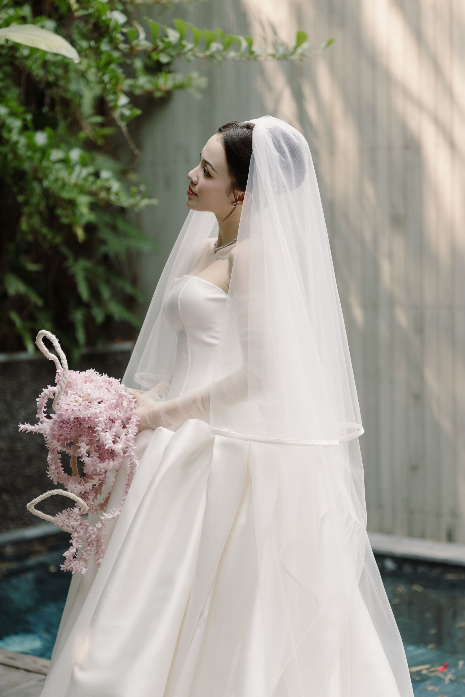 Elegant bride in strapless white gown and veil holding pink floral bouquet beside garden pool.