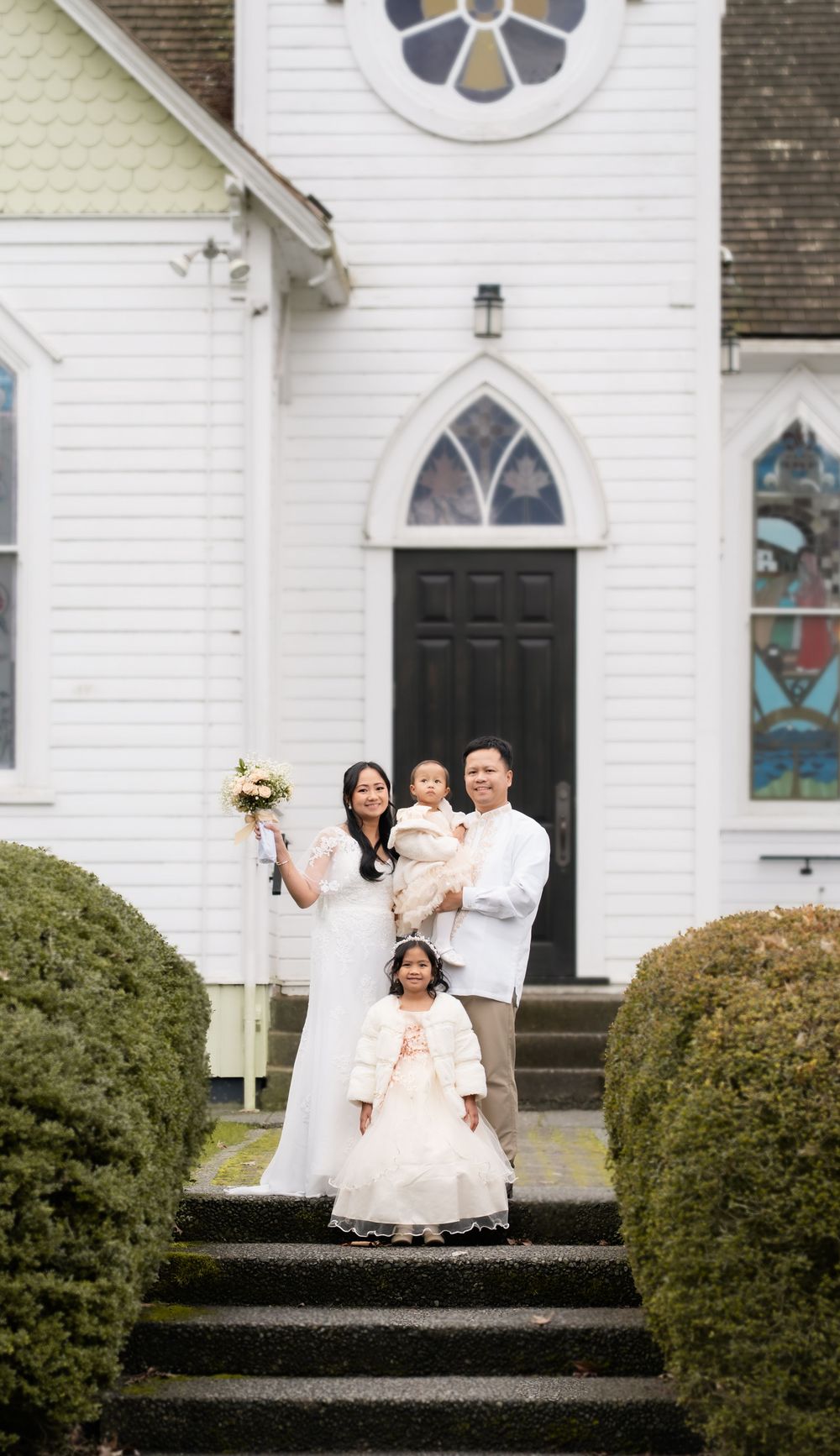 Family in white wedding attire poses on church steps with stained glass windows and manicured hedges.