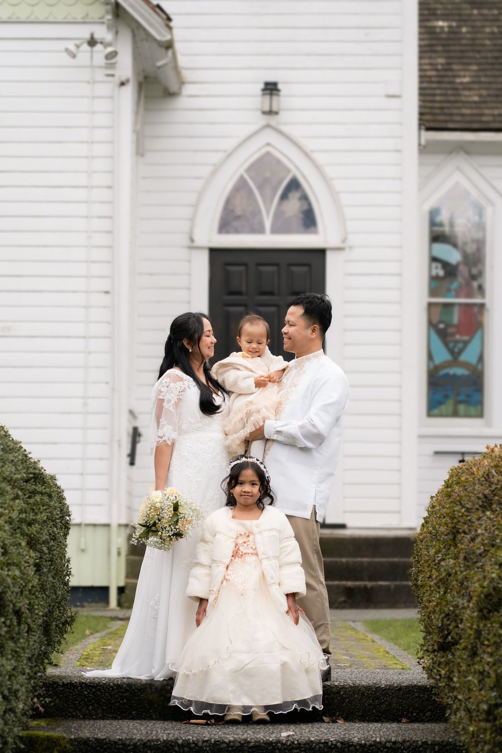 Family poses in wedding attire outside white church, parents holding children in elegant white dresses on steps.