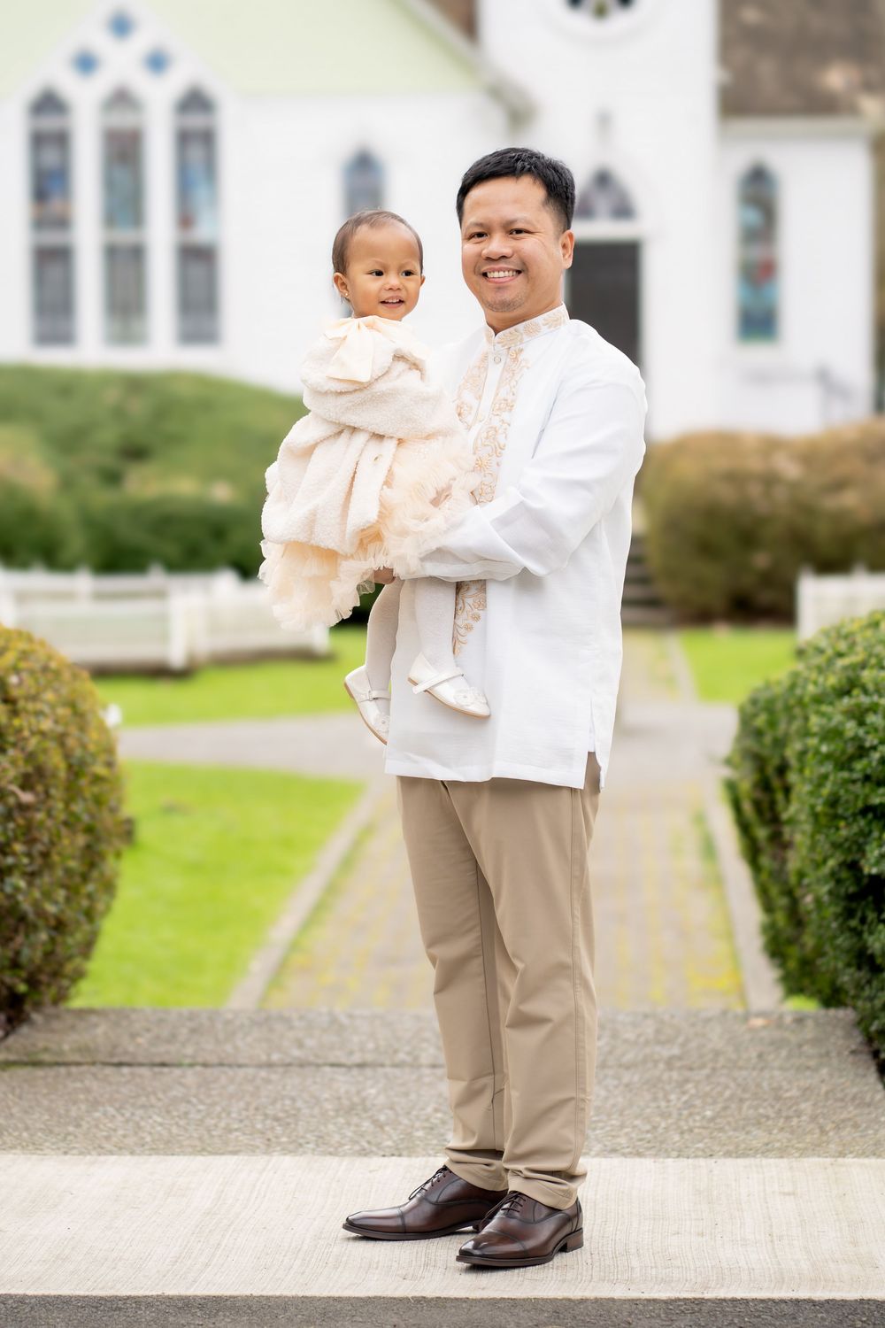 Filipino man in barong holds baby girl in white dress outside a church on a sunny day.