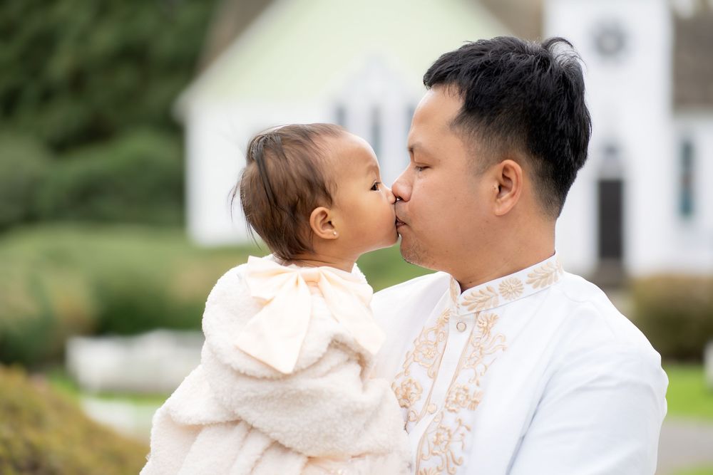 A father tenderly kisses his baby girl outdoors, both dressed in white, with a house and greenery in the background.