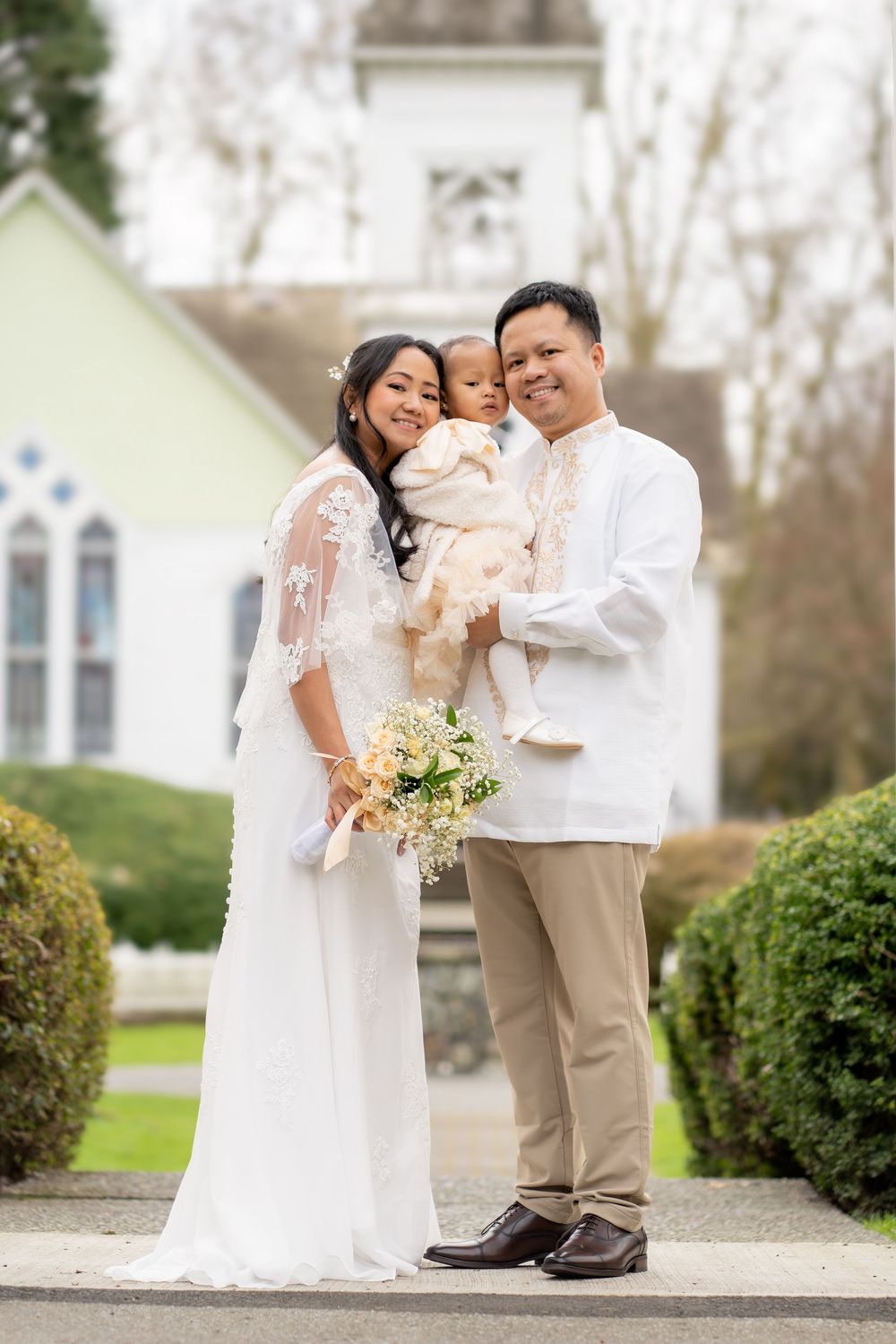 Filipino family in white formal attire poses outside a church, parents holding baby after a baptism ceremony.