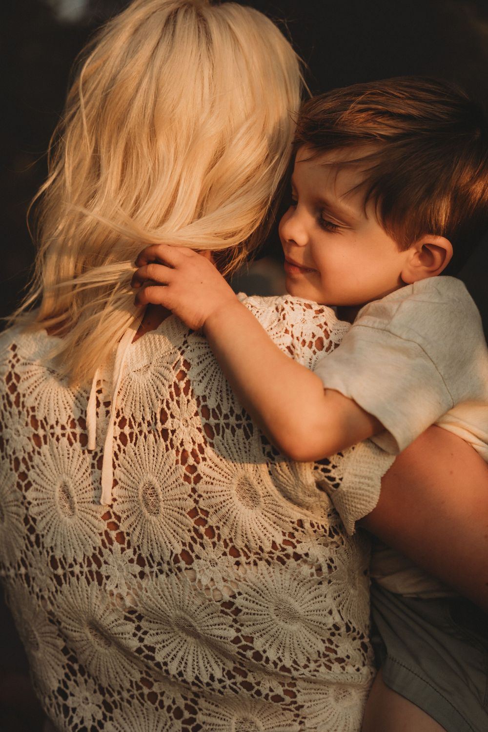 A young child lovingly embraces a blonde woman in lace top, bathed in warm golden sunlight.