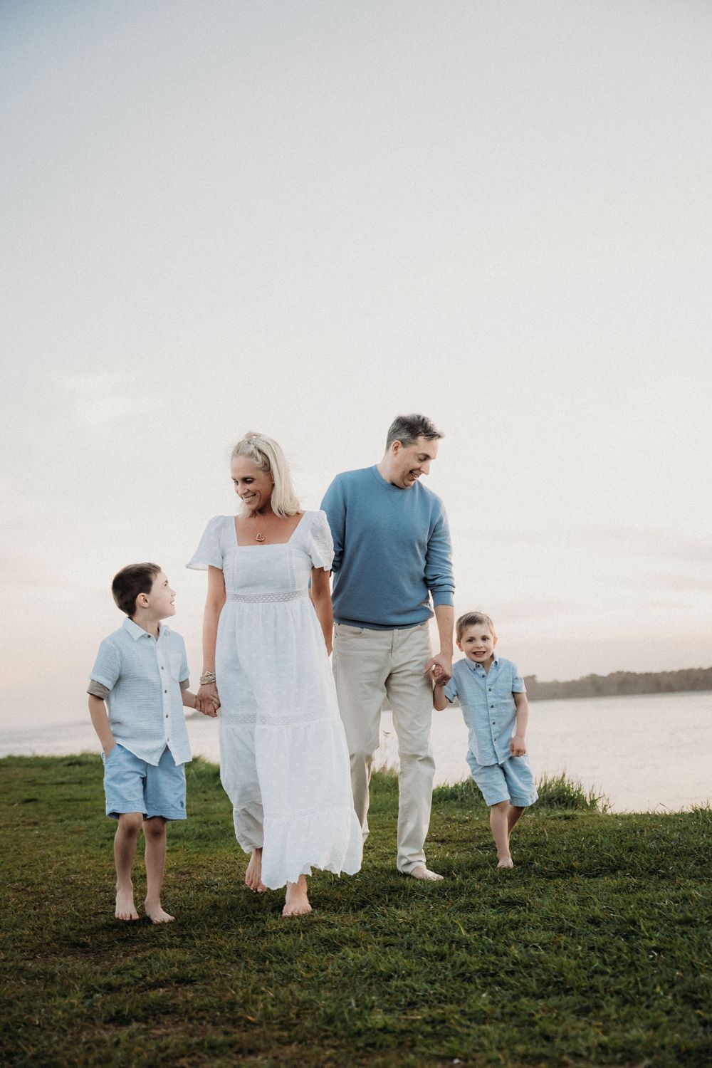 Family of four walking hand-in-hand by a lakeside at dusk, wearing coordinated blue and white outfits.