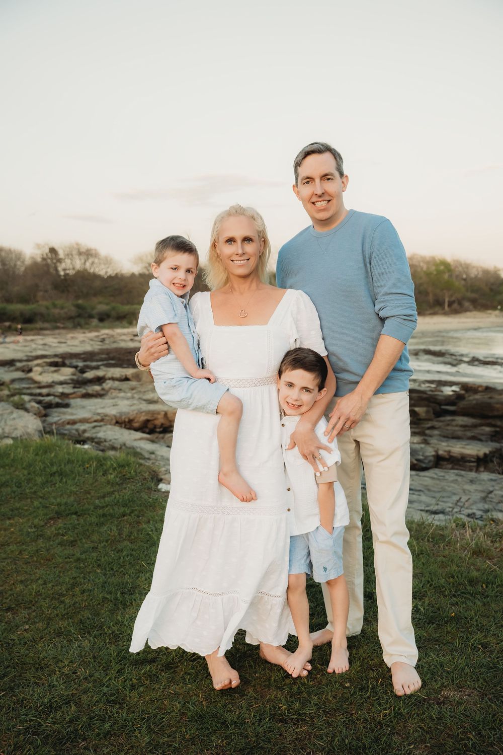 Family of four posing outdoors near rocky coastline at sunset, wearing coordinated blue and white outfits.