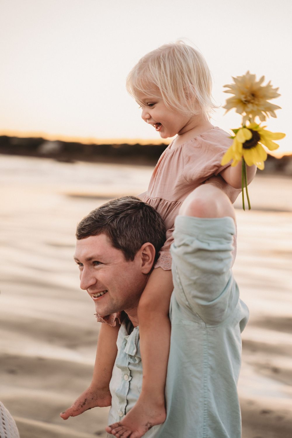 A smiling father carries his laughing blonde toddler on his shoulders at a sunset beach, as she holds a bright yellow sunflower.