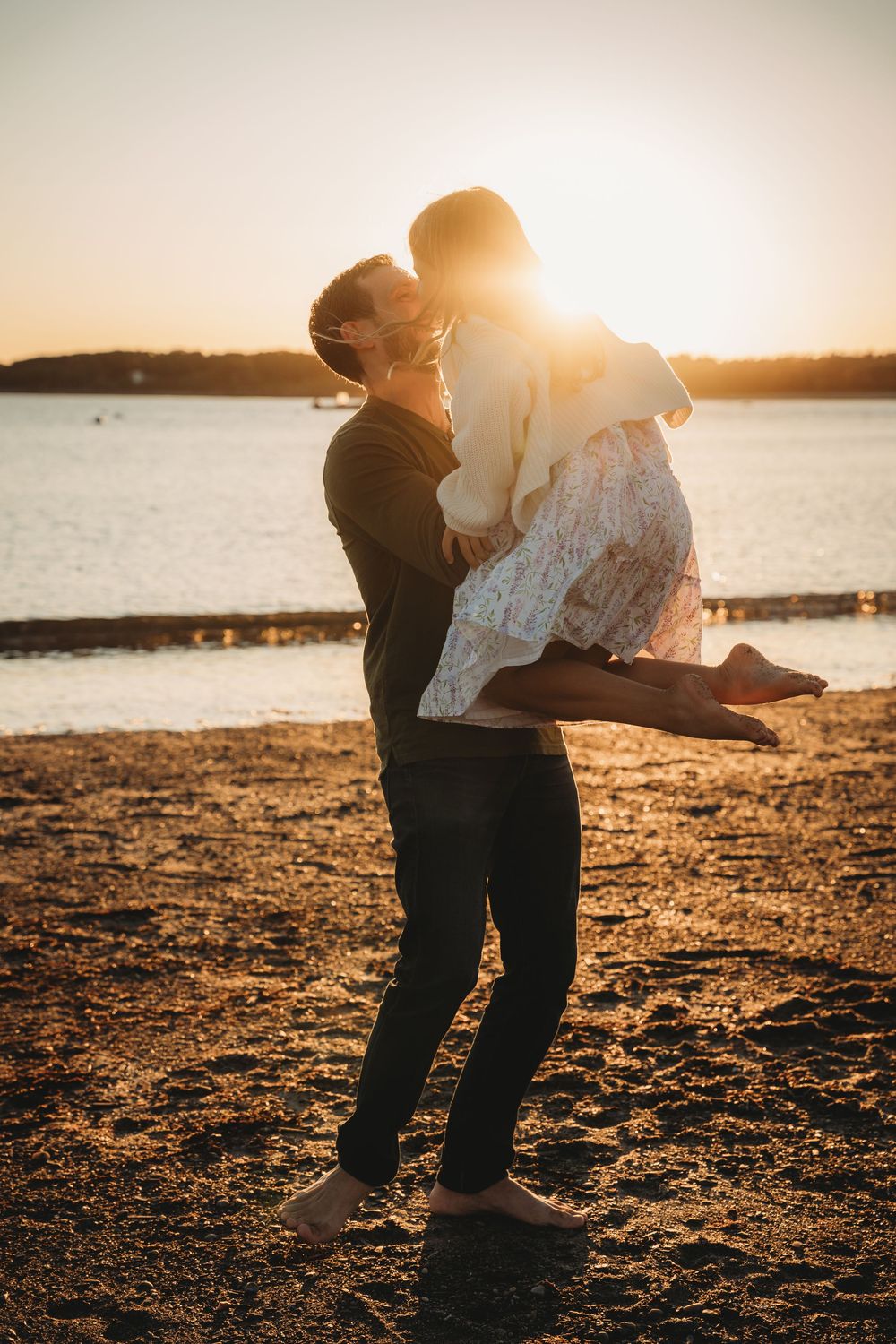 A couple shares a romantic kiss at sunset on a sandy beach, as the man lifts the woman in his arms.