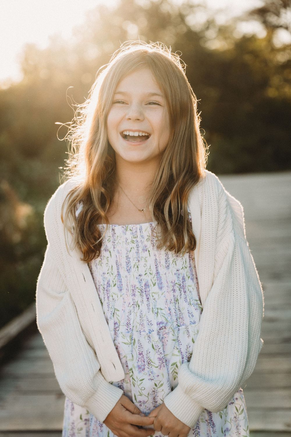 A laughing young girl with long brown hair wearing a floral dress and white cardigan, bathed in golden sunset light outdoors.