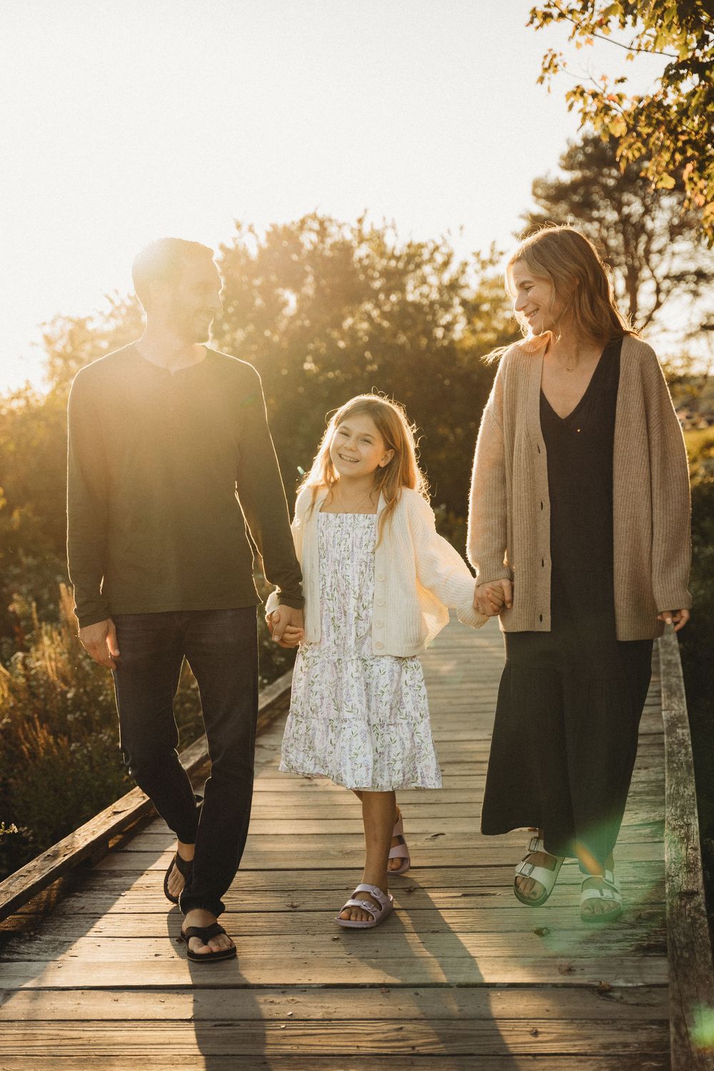 Family of three holding hands walking on a wooden boardwalk at golden hour sunset.