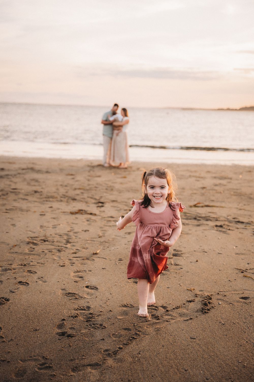 A joyful little girl in a pink dress runs on a sandy beach at sunset while her parents embrace by the ocean.