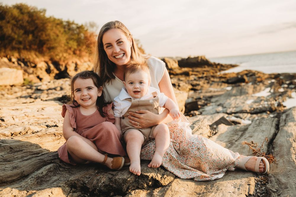 Mother sitting on rocky beach with two young children, smiling warmly at sunset in a coastal outdoor family portrait.