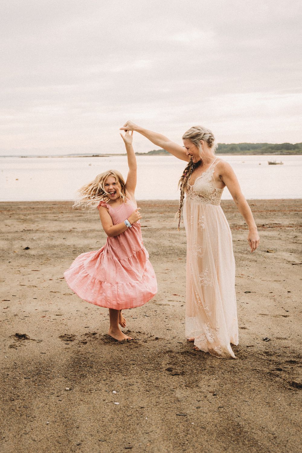 Woman in white gown twirling young girl in pink dress on sandy beach, joyful mother-daughter dance by the ocean.