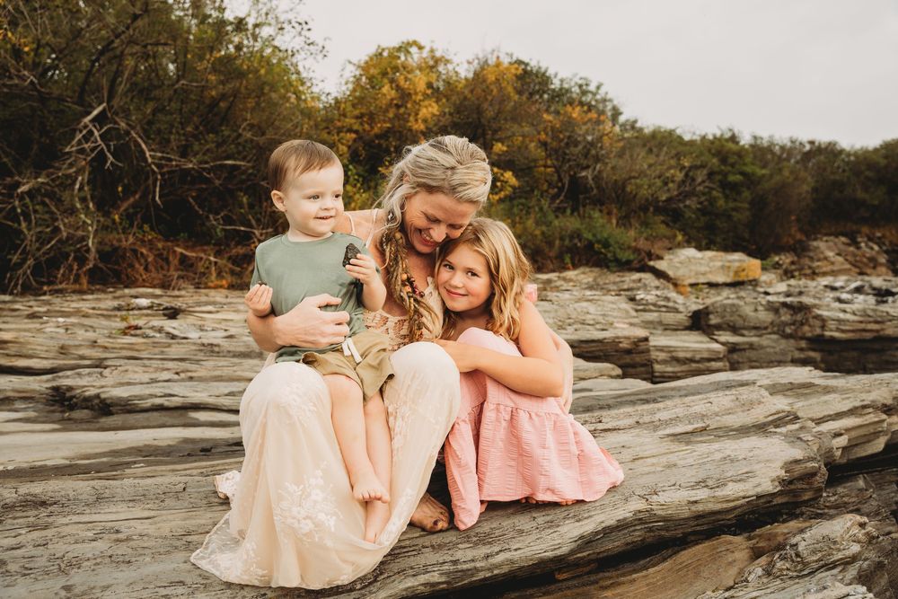 A mother sits on rocks outdoors, lovingly embracing her two young children in a warm family portrait.