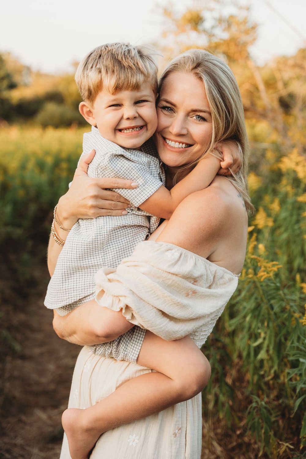 A smiling blonde woman in an off-shoulder dress holds a laughing young boy in a sunny outdoor field.