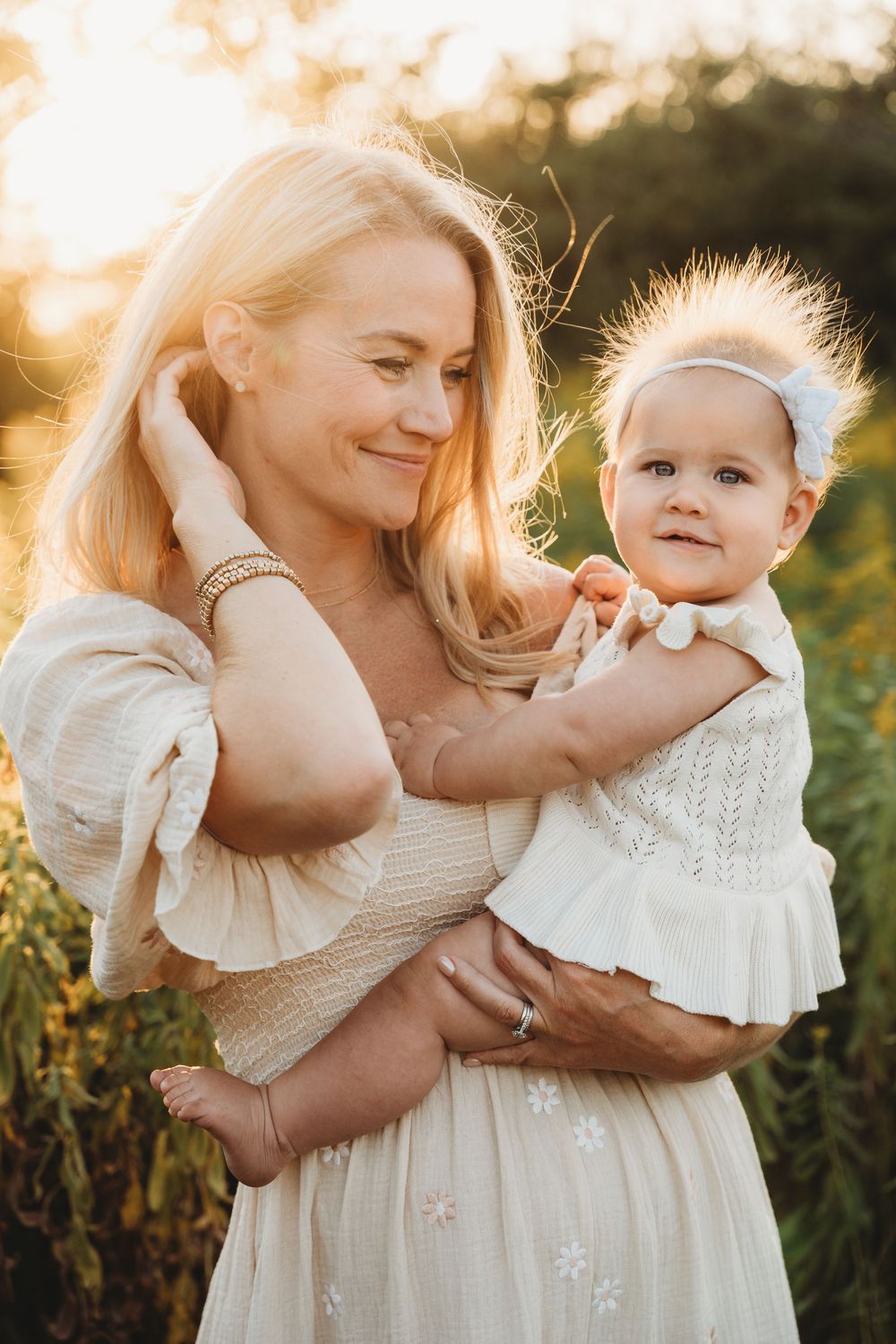 A woman in a white floral dress holds a baby girl in a white outfit during a golden sunset outdoors.