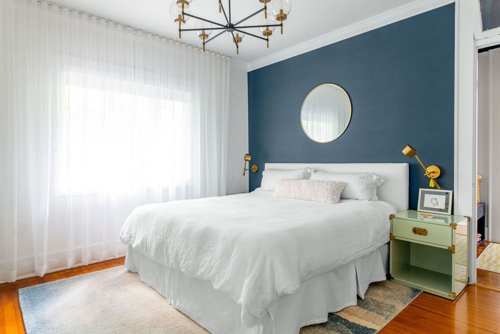 Cozy bedroom with blue accent wall, round mirror, white bedding, and gold chandelier.