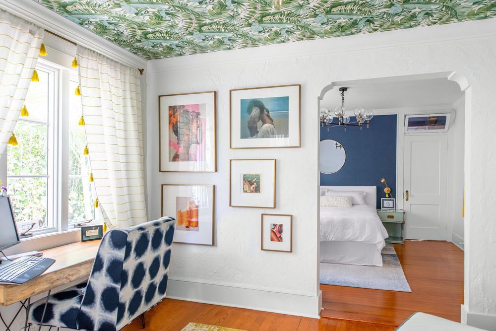 Stylish home office with floral wallpaper ceiling, gallery wall art, ikat chair, and view into blue bedroom.