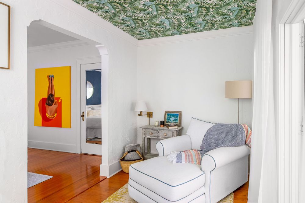Cozy reading nook with white armchair, ottoman, floral wallpapered ceiling, hardwood floors, and colorful artwork visible through archway.