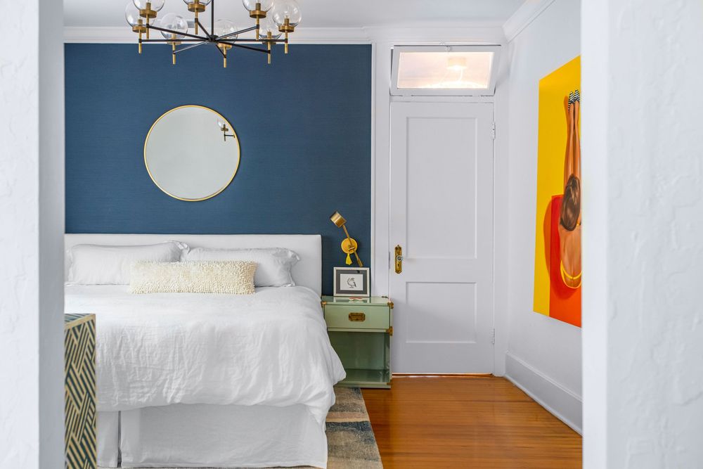 Cozy bedroom with navy blue accent wall, round mirror, white bedding, green nightstand, and vibrant guitar artwork near white door.