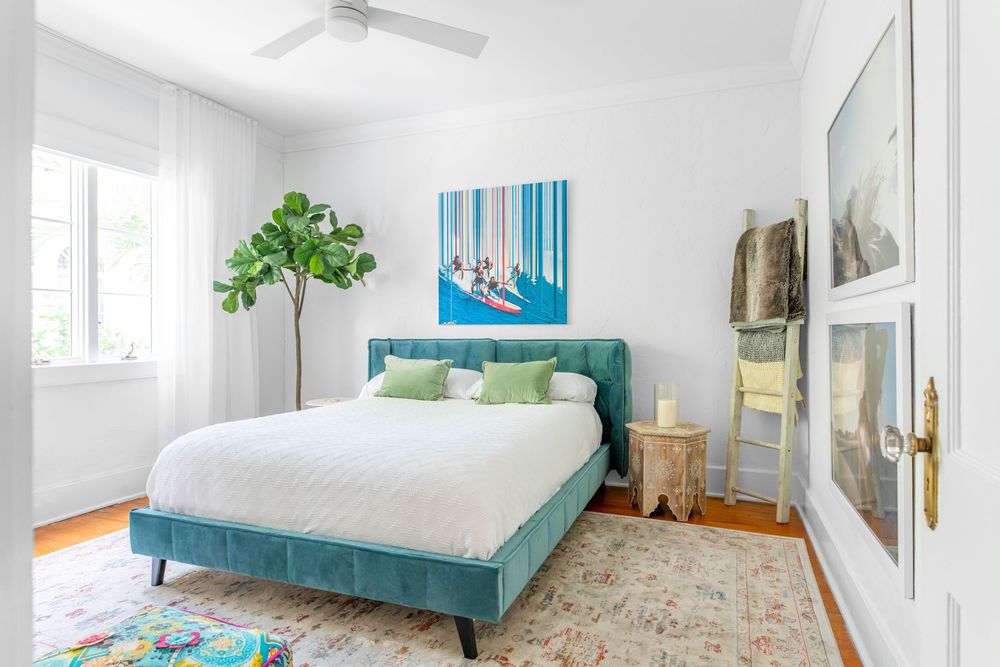 Bright bedroom with teal velvet bed, colorful abstract artwork, fiddle leaf fig tree, and hardwood floors.