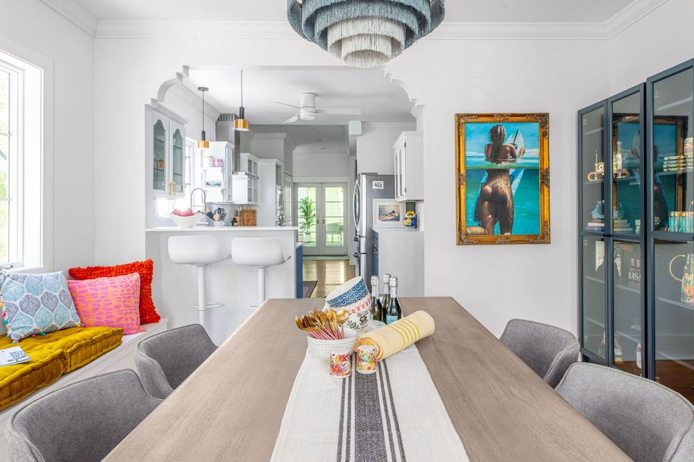 Modern dining room with gray chairs, wood table, colorful artwork, and open kitchen view in a bright white home.