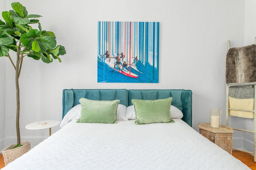 Bright bedroom with teal upholstered bed, green pillows, surfing wall art, and a fiddle leaf fig tree.