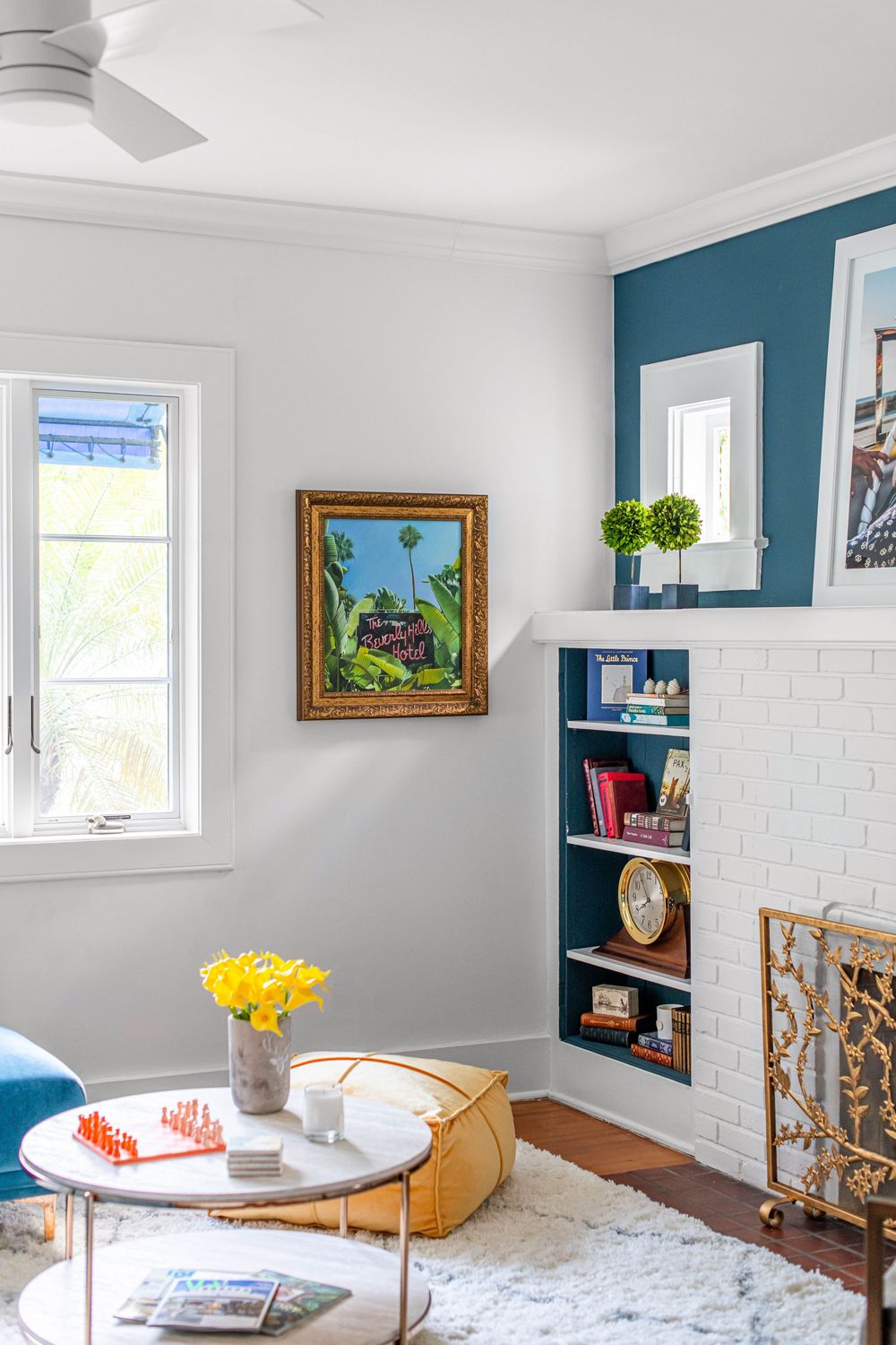 Bright living room with teal accent wall, white brick fireplace, built-in bookshelf, gold-framed art, and a round coffee table.