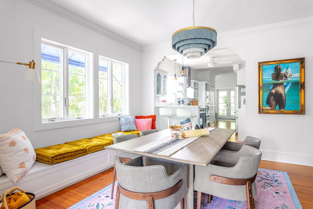 Bright dining room with long table, gray chairs, built-in window bench with yellow cushion, colorful rug, and modern pendant light.