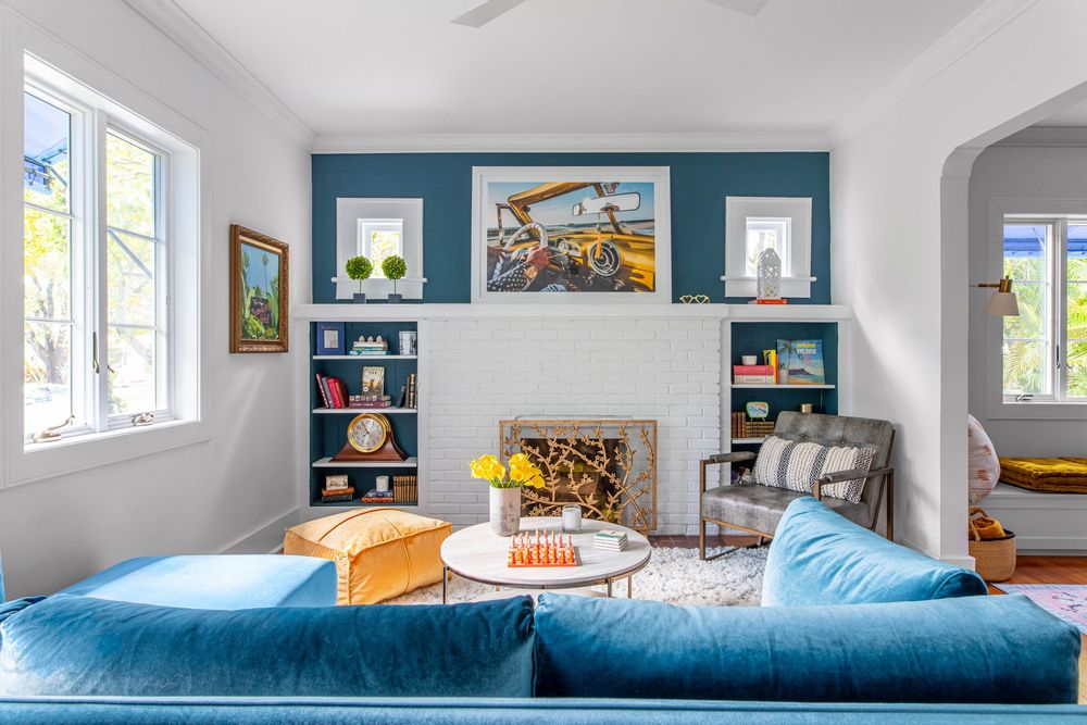 Bright living room with blue velvet sofas, white fireplace, teal accent wall, built-in shelves, and colorful artwork.